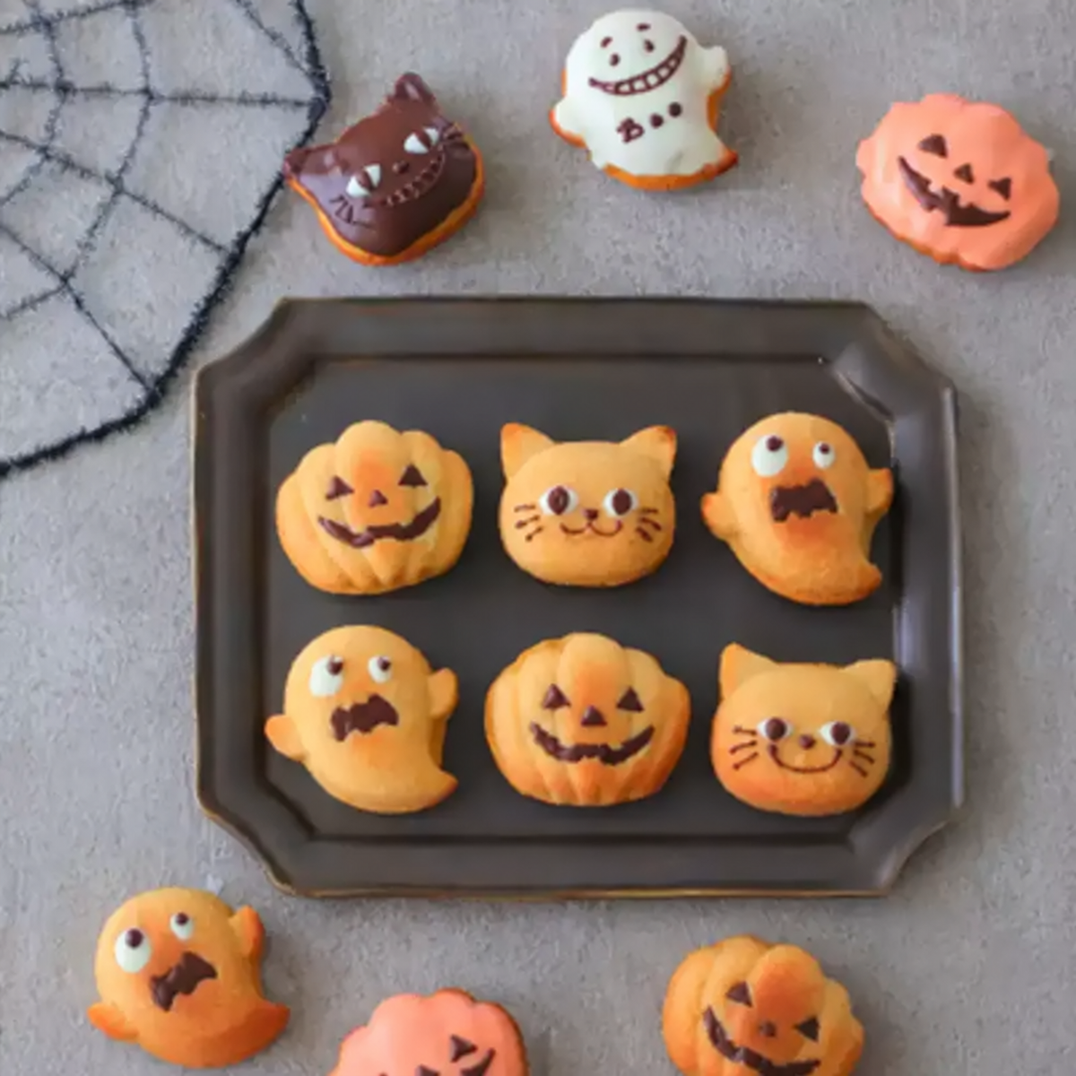 Golden pumpkin, ghost and cat mini cakes with chocolate faces arranged on a dark baking tray.