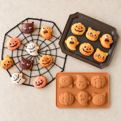 Orange six-cavity Halloween silicone mould beside decorated pumpkin, ghost and cat cakes on a spiderweb mat and baking tray.